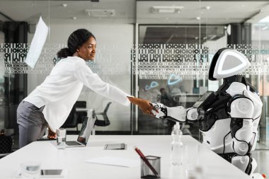 Gülümseyen Afrikalı Amerikalı iş kadını ofiste robotla el sıkışıyor.