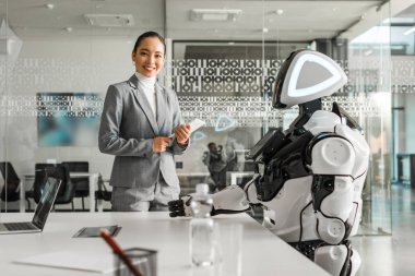 Toplantı odasında robotun yanında dururken elinde dijital tablet tutan gülümseyen Asyalı kadın.