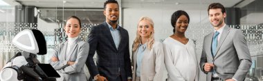 panoramic shot of smiling multicultural businesspeople looking at camera while standing near robot clipart