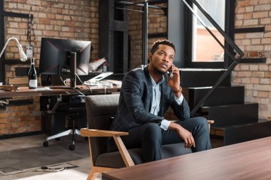 Düşünceli afro-amerikan iş adamı koltuğunda oturup akıllı telefondan konuşurken yukarı bakıyor.