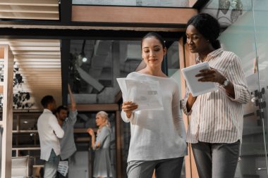 Dijital tabletli ve belgeli genç Afrikalı Amerikalı ve Asyalı iş kadınları arka planda çok kültürlü meslektaşlarıyla birlikte büroda duruyorlar.