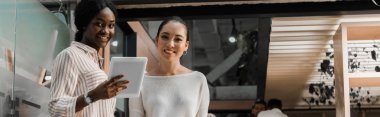 Sayısal tablet tutan Afro-Amerikan iş kadınının meslektaşının yanında dururken çekilmiş panoramik bir fotoğrafı.