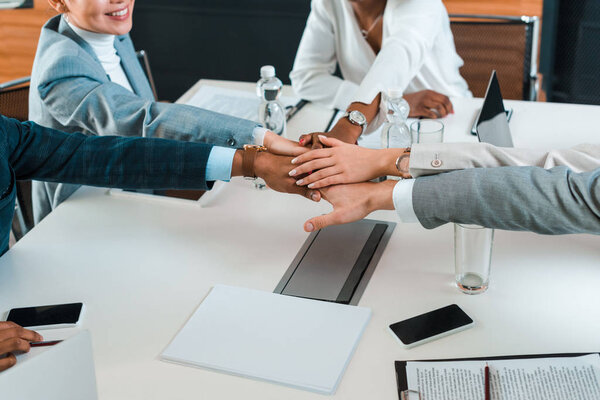 partial view of young multicultural businesspeople holding joined hands while sitting at desk in conference hall