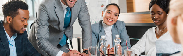 обрезанный вид молодого бизнесмена, стоящего рядом с улыбающимися мультикультурными коллегами, сидящими за столом в конференц-зале, панорамный снимок
