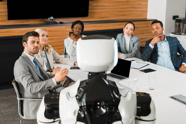 selective focus of young multicultural businesspeople looking at robot while sitting in conference hall