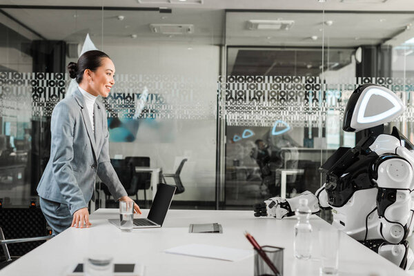 smiling asian businesswoman looking at robot sitting at desk in office