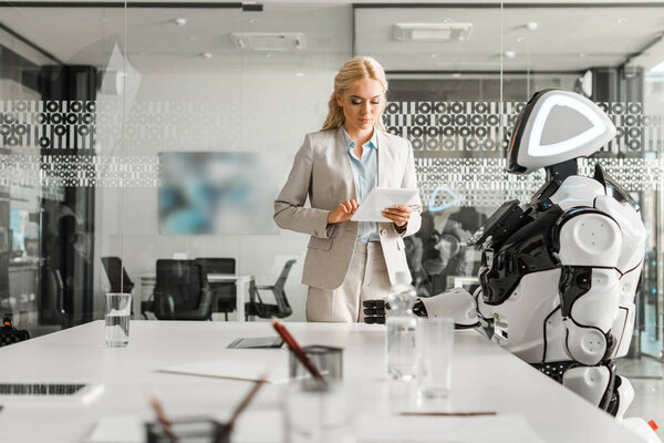 attractive businesswoman using digital tablet while standing near robot in meeting room