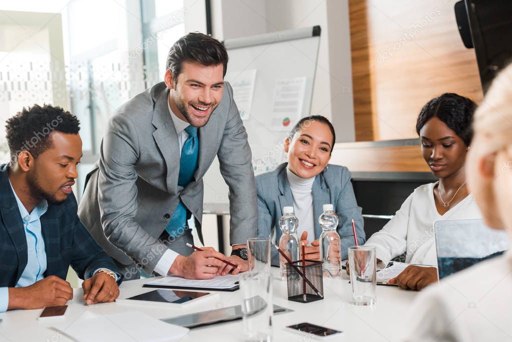 selective focus of cheerful businessman holding pencil while standing near multicultural colleagues sitting at desk in conference hall