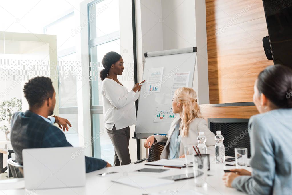 pregnant african american businesswoman pointing with pen at flipchart near multicultural colleagues