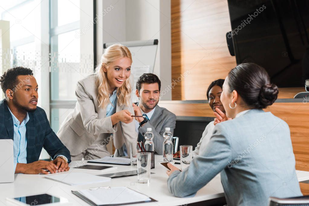 young smiling businesswoman pointing with pencil near multicultural colleagues sitting at desk in conference hall