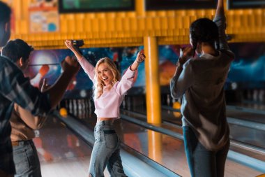 excited blonde girl showing winner gesture while standing near bowling alley with multicultural friends clipart