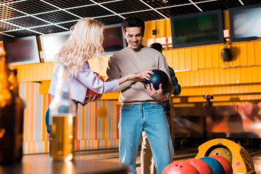 Sarışın kız, gülümseyen erkek arkadaşının ellerinde bowling topuna dokunuyor.