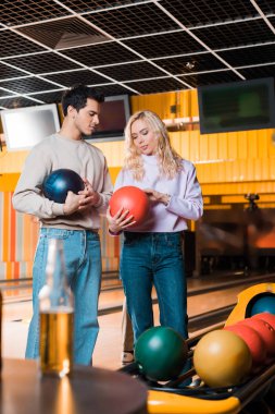 Genç adam ve kadın bowling kulübünde bowling toplarını tutarken konuşuyorlar.