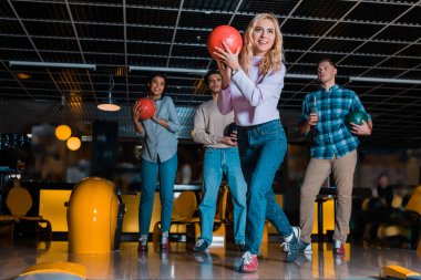 smiling blonde girl throwing bowling ball near multicultural friends clipart