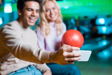selective focus of smiling girl holding bowling ball while cheerful boyfriend taking selfie on smartphone clipart