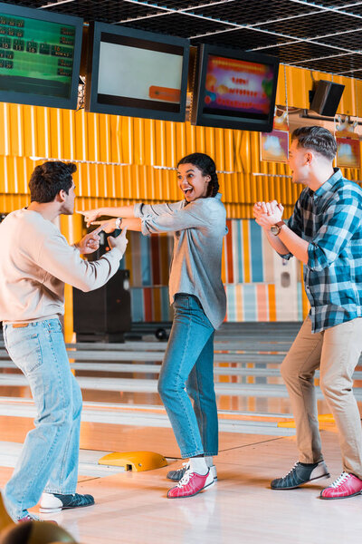 happy african american girl pointing with fingers while standing near excited multicultural friends in bowling club