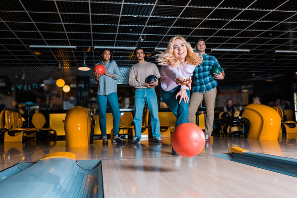 smiling blonde girl throwing bowling ball on skittle alley near multicultural friends