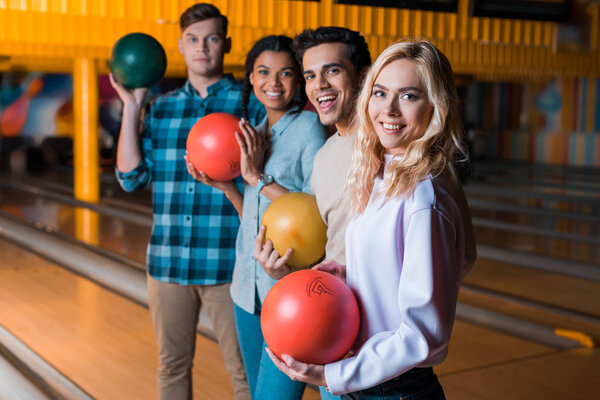 happy multicultural friends holding bowling balls and looking at camera while standing in bowling club