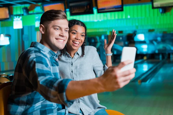陽気な異人種間のカップルがボウリングクラブに座っている間にスマートフォンで自撮り — ストック写真
