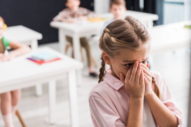 selective focus of upset schoolkid crying near classmates, bullying concept  clipart