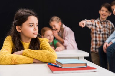 selective focus of upset schoolgirl near classmates gossiping and pointing with finger isolated on black, bullying concept  clipart