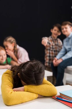 Seçici odak noktası, sınıf arkadaşlarının yanında yüzünü örten, dedikodu yapan ve siyah, zorbalık kavramına izole edilmiş parmakla işaret eden üzgün kız. 