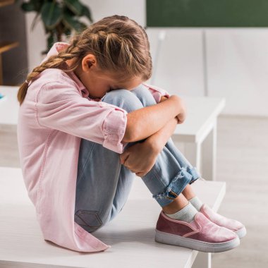 bullied schoolgirl covering face while sitting on desk in classroom  clipart