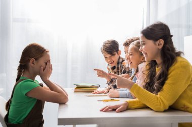 Zalim okul çocukları parmaklarıyla işaret ediyor ve okulda ağlarken yüzünü örten zorba kızın yanında gülüyorlar. 
