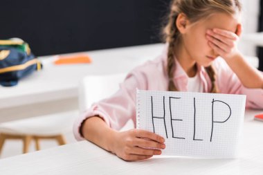 selective focus of paper with help lettering in hand of upset schoolgirl covering eyes, bullying concept clipart