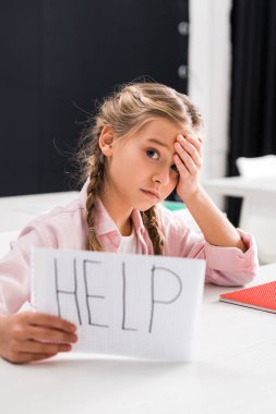 selective focus of upset schoolgirl holding paper with help lettering, bullying concept clipart