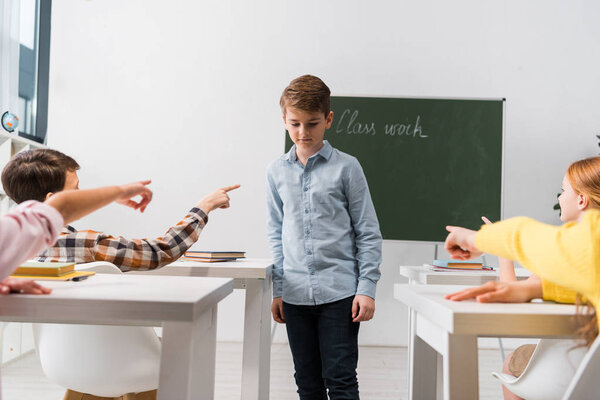 selective focus of classmates pointing with fingers at frustrated schoolboy covering face, bullying concept 