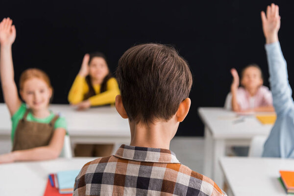 back view of schoolboy standing near classmates with raised hands isolated on black 