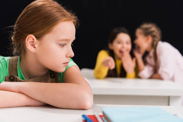 selective focus of upset schoolkid sitting near classmates laughing and gossiping in classroom isolated on black, bullying concept 