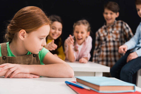selective focus of upset schoolkid sitting near classmates laughing and pointing with fingers isolated on black, bullying concept 