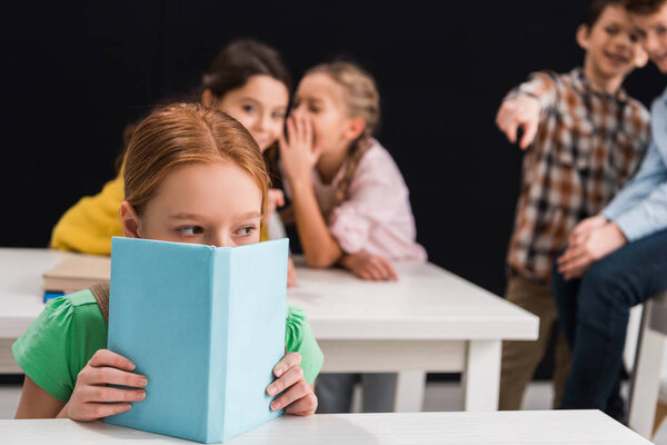 selective focus of upset schoolkid covering face with book near classmates gossiping and pointing with finger isolated on black, bullying concept 