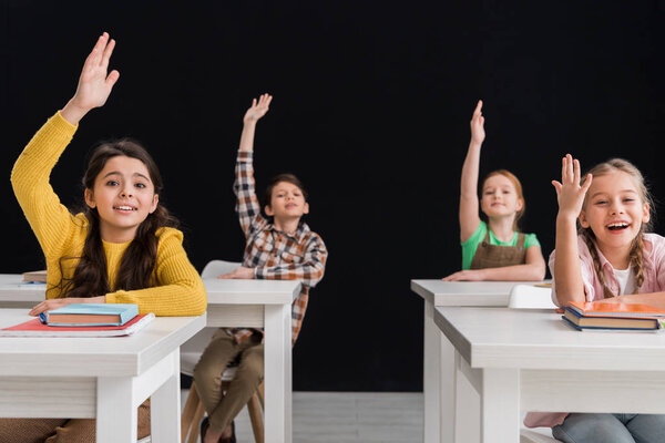 selective focus of happy schoolchildren with raised hands isolated on black