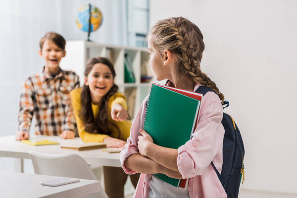 selective focus of bullied schoolgirl holding notebooks near cruel schoolkids 