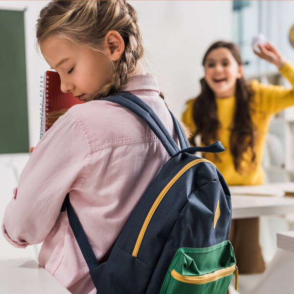 selective focus of bullied schoolgirl with backpack holding notebooks near cruel schoolkid