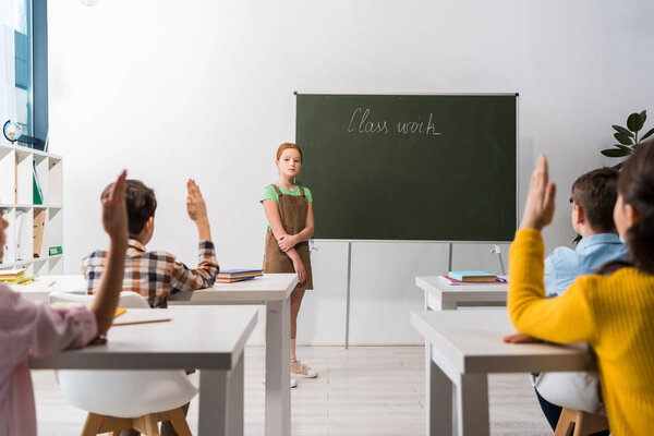 selective focus of schoolkgirl standing near chalkboard with class work lettering and classmates with raised hands 