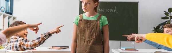 panoramic shot of cruel classmates pointing with fingers at bullied schoolgirl in classroom 