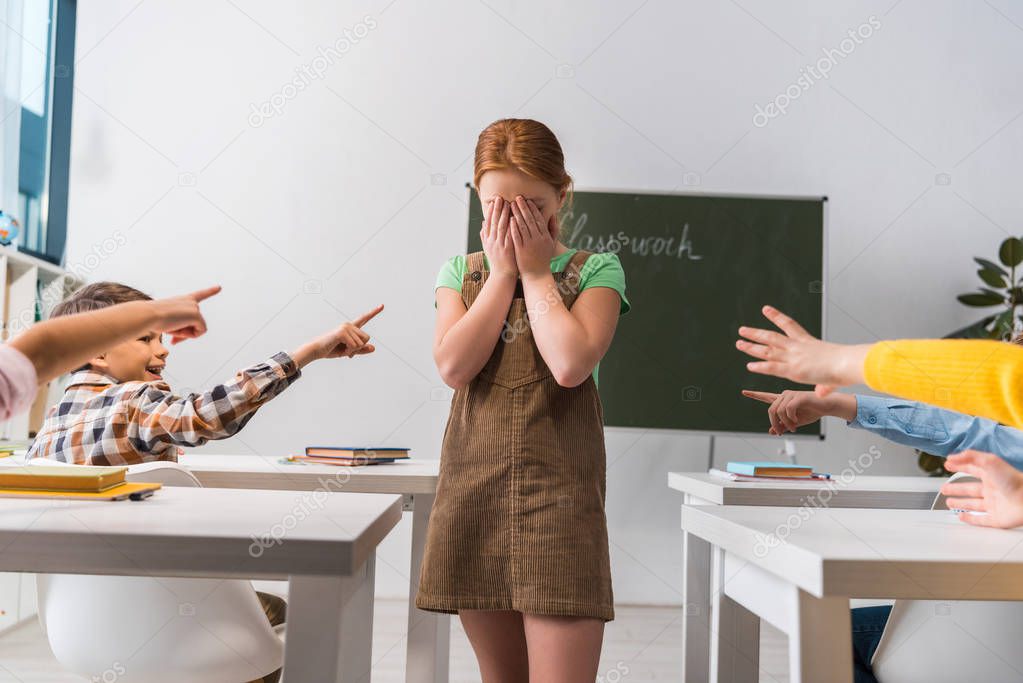 cruel schoolboy pointing with finger at frustrated classmate near schoolkids in classroom, bullying concept 