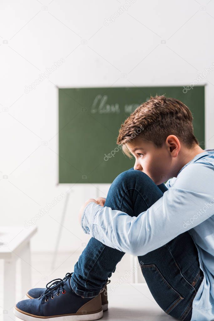 bullied schoolboy sitting on table in classroom 
