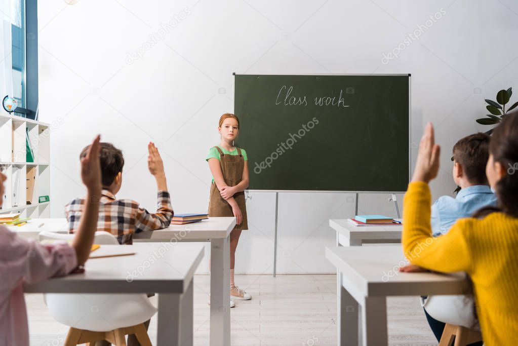selective focus of schoolkgirl standing near chalkboard with class work lettering and classmates with raised hands 