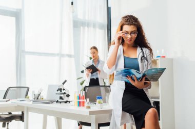 Gözlüklü ve beyaz önlüklü seksi hemşirenin laboratuvarda kadının yanında fen dergisi okuduğu seçici odak noktası. 