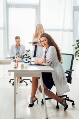 Laboratuvarda beyaz önlüklü kadınların yanında otururken gözlük tutan seksi hemşire. 