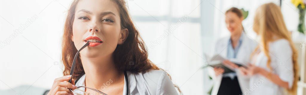 panoramic shot of sexy nurse holding glasses near lips in clinic 