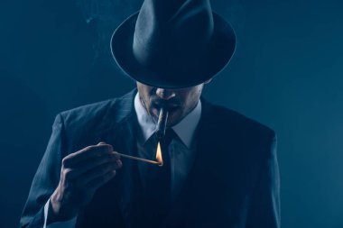 Mafioso with covered eyes with felt hat lighting cigar on dark blue background clipart