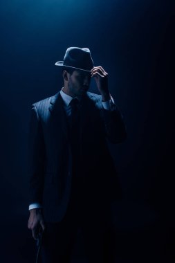 Gangster touching felt hat and holding gun on dark background clipart