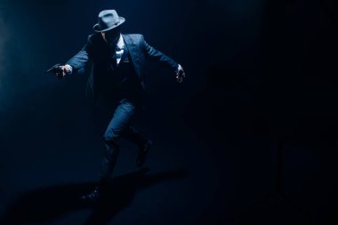 Geniş elli bir gangster tabancayı hedefliyor ve karanlık arka planda koşuyor.