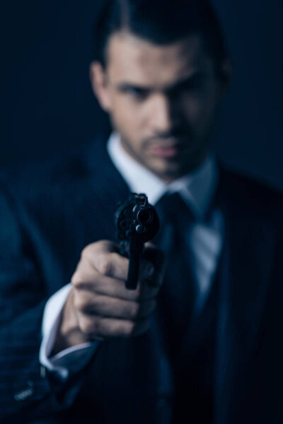 Selective focus of gangster aiming gun and looking at camera on dark background